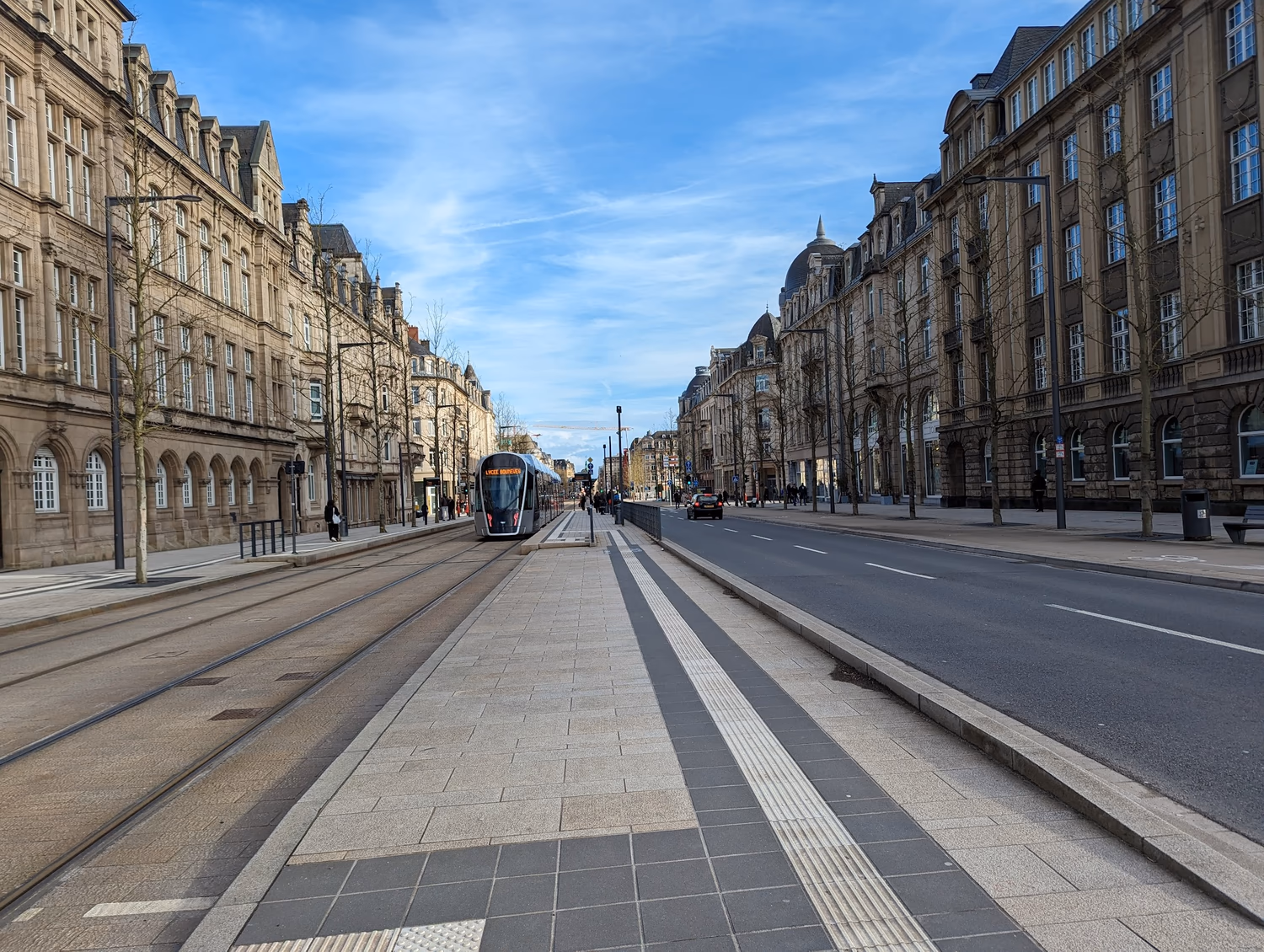 A photo I took standing in the middle of one of the main streets in Luxembourg. On either side are shops of about 4 floors, which are maybe 2/5 the height of the width of the street. There are quite wide pavements, and on the left two tram lanes and a tram in shot, and more pavement and tramstops in the centre. To the right are two reasonably narrow road lanes and to its right a cycle path. The buildings and street (although not the road) are yellow-grey and well-kept, with trees, benches, and bins.